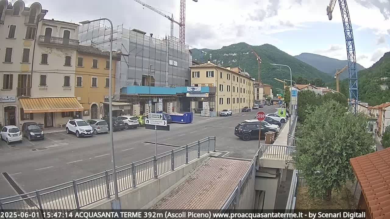 A street scene in a town nestled in mountains under a partly cloudy sky shows several cars parked along the sides of a street, buildings under construction, and a crane in the background.