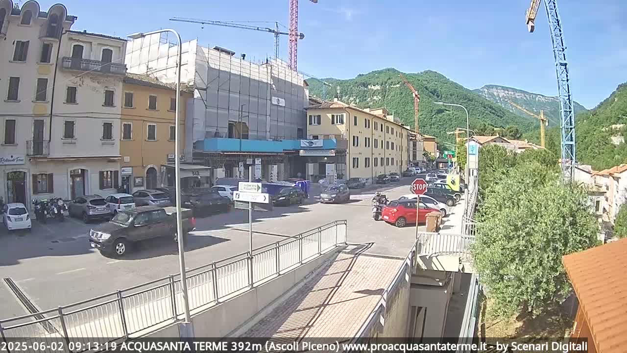 A sunny day reveals a street scene in a town nestled in a valley, with buildings under construction and several cars parked along the roadside.