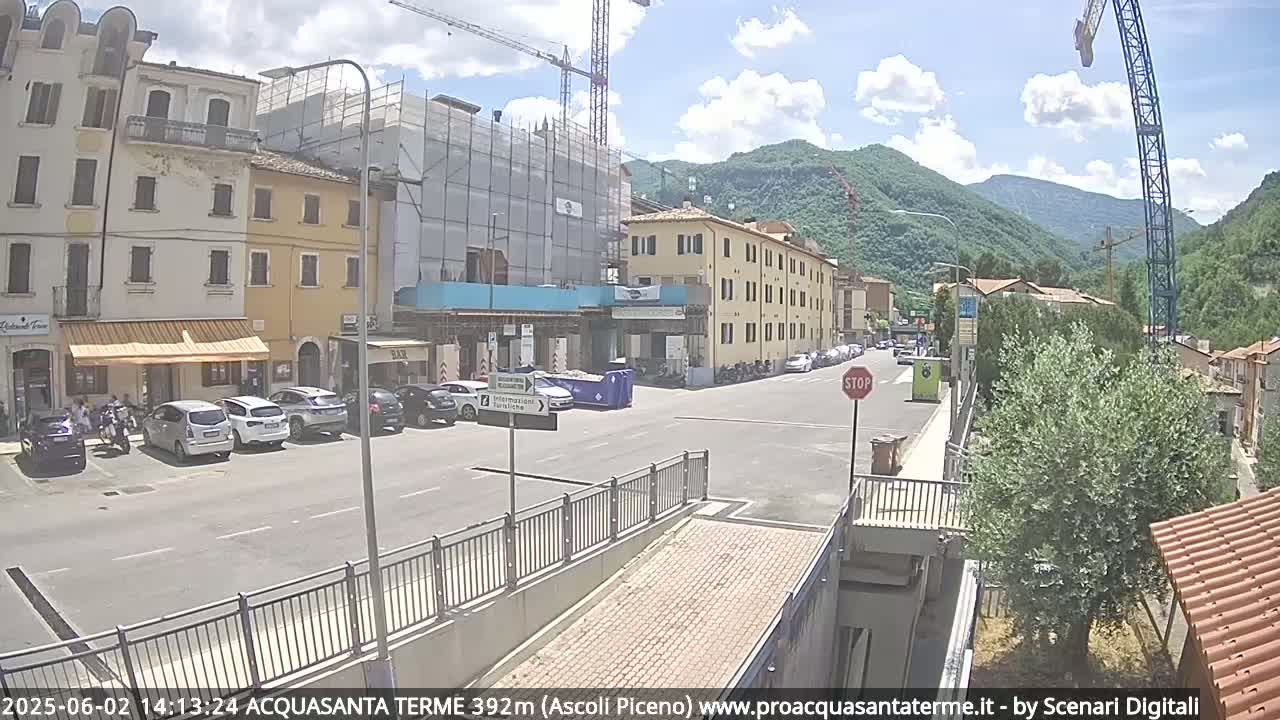 A sunny day reveals a town square with parked cars, buildings under construction, and mountains in the background.