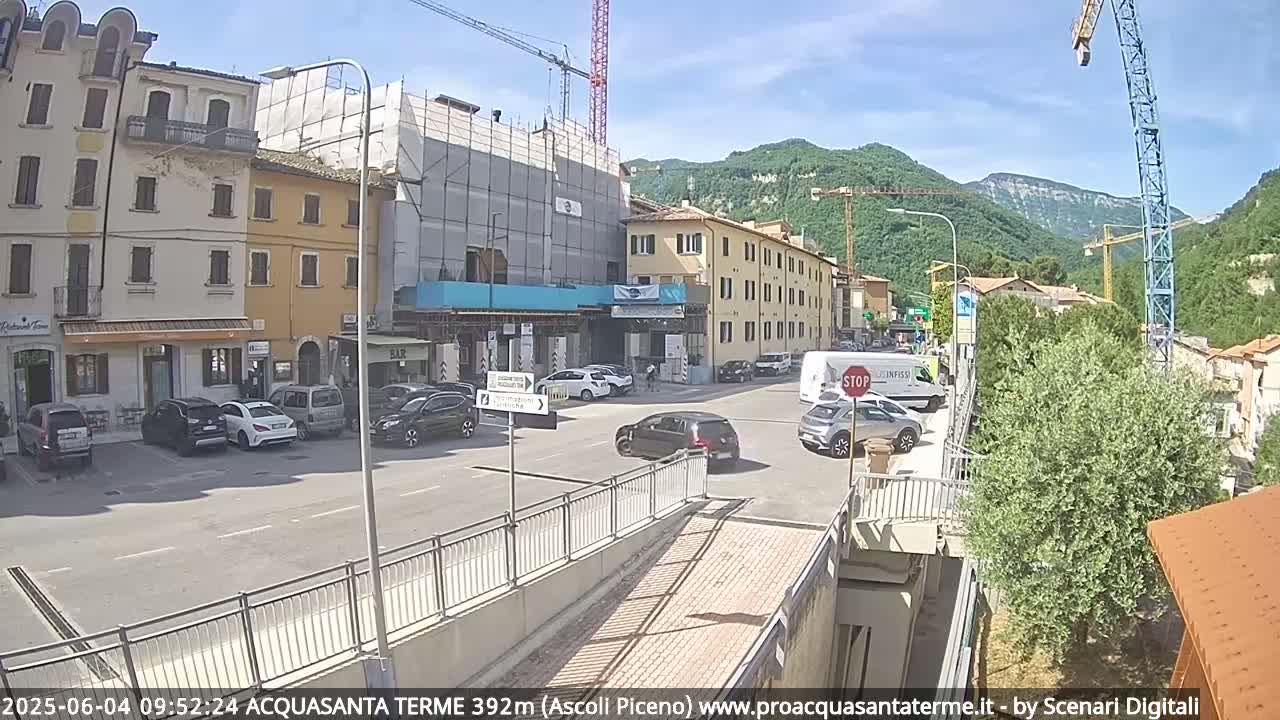 A sunny day reveals a town square with parked cars, a building under construction, and several other buildings, all situated against a backdrop of green hills.