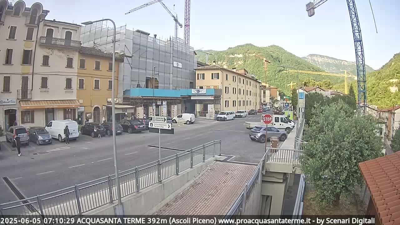 A street scene in a town shows several buildings, some under construction, with cars parked along the side of the road and a few cars driving on a sunny day.