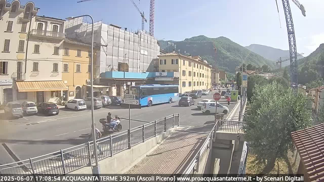 A sunny day in a town nestled in a valley, with a blue bus and several cars parked along the side of a street near a building under construction.