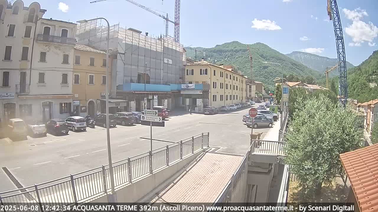 A sunny day reveals a town square with parked cars, buildings under construction, and mountains in the background.