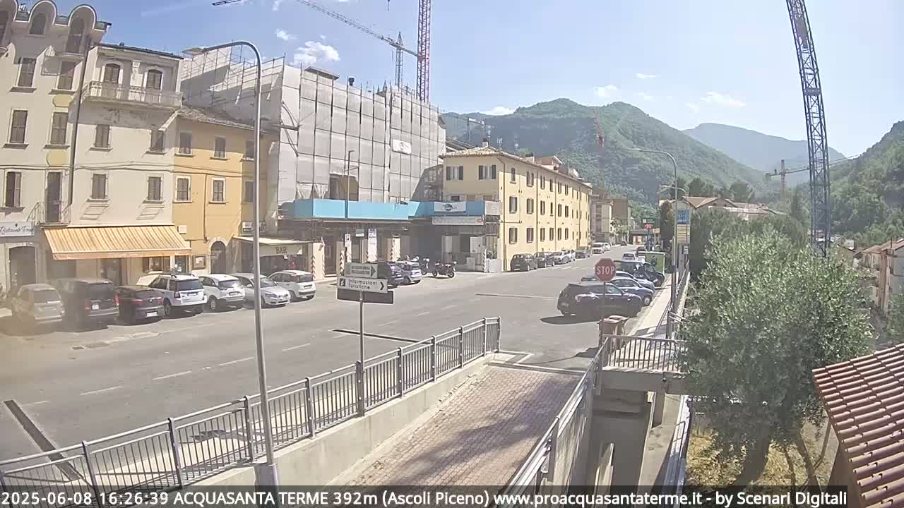 A sunny street scene in a town nestled in a valley, showing buildings, parked cars, and a construction site.