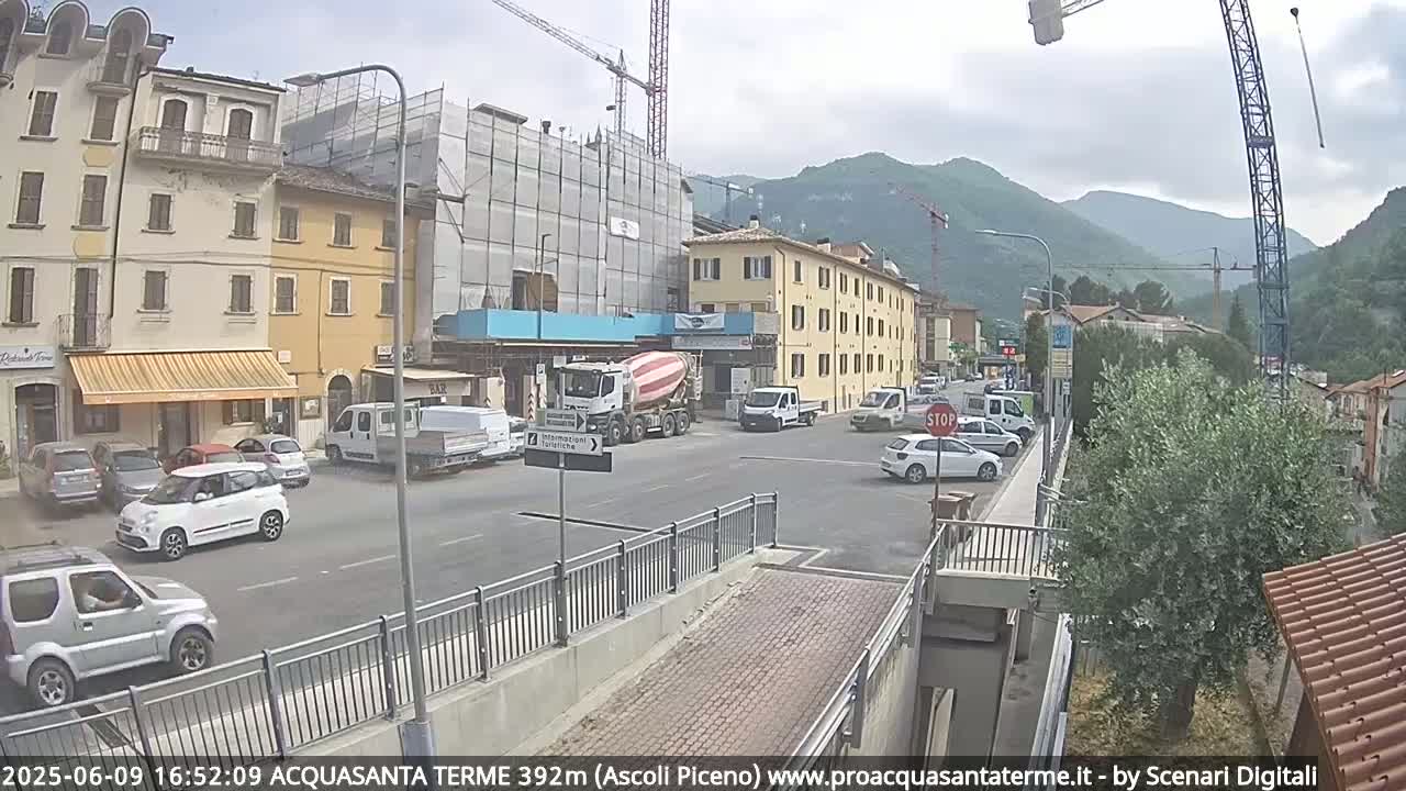 A street scene in a town shows several cars parked and driving on a road near buildings under construction, with mountains in the background under an overcast sky.