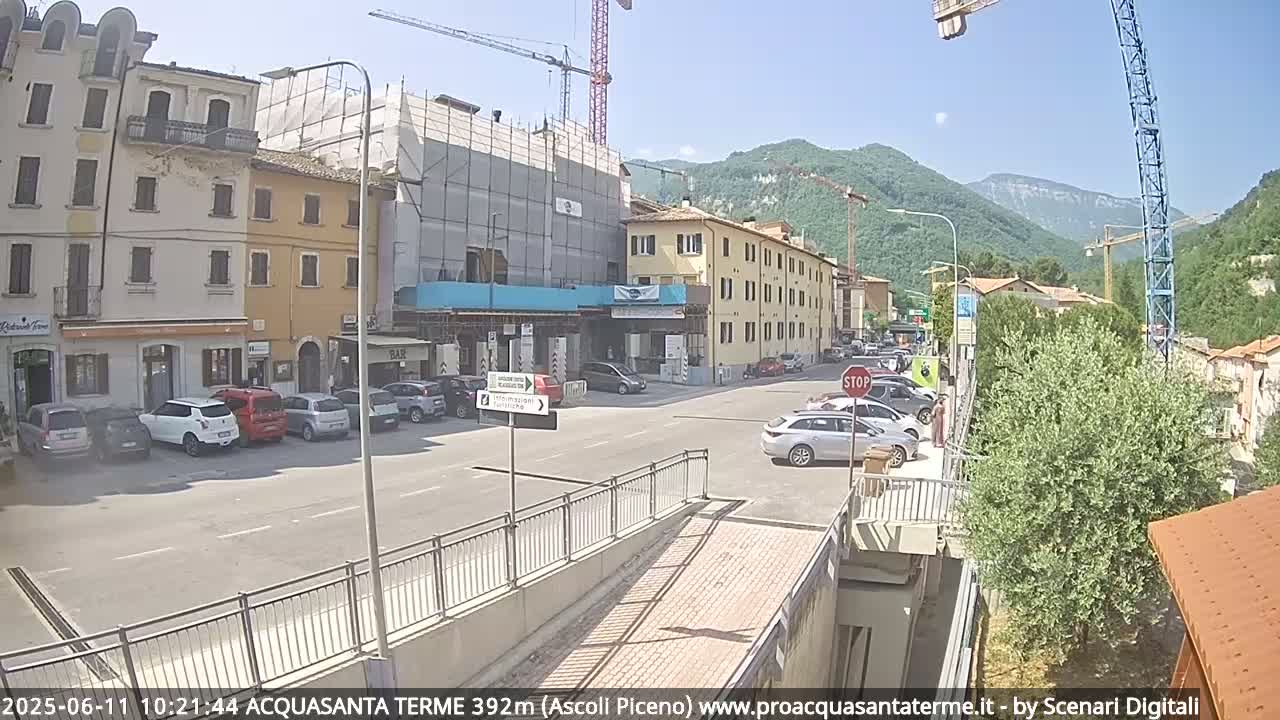 A sunny town square with parked cars, several buildings including one under construction, and mountains in the background.