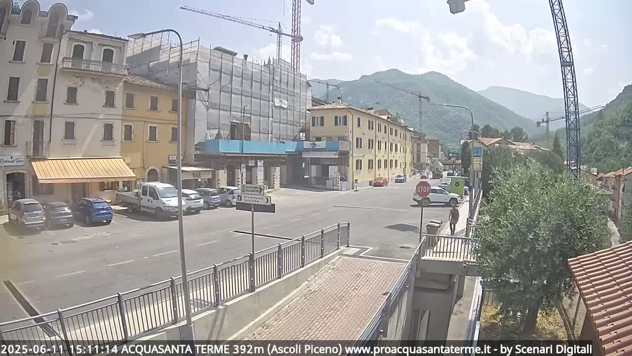 A sunny day in a town square with several parked cars, buildings under construction, and mountains in the background.