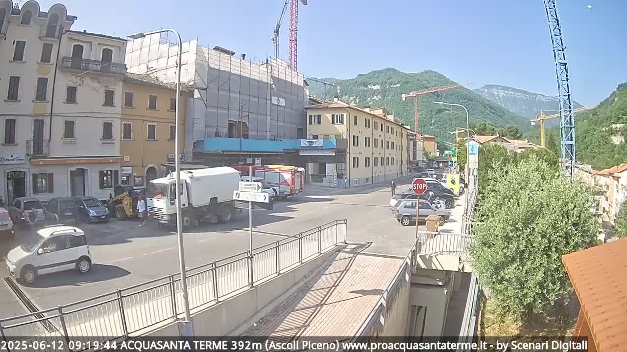 A sunny day reveals a street scene with parked cars, construction, and buildings, backed by green hills and cranes.