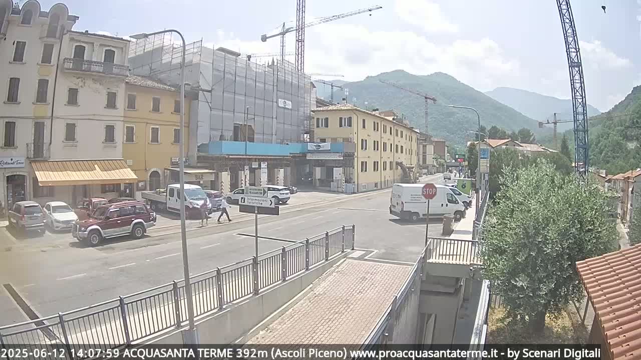 A street scene shows several cars parked near buildings under construction on a sunny day, with mountains in the background.