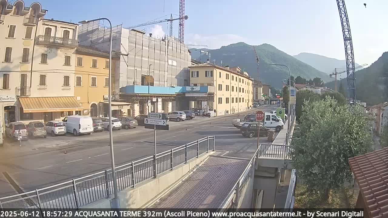A sunny day in a town with buildings under construction, cars parked along the street, and mountains in the background.