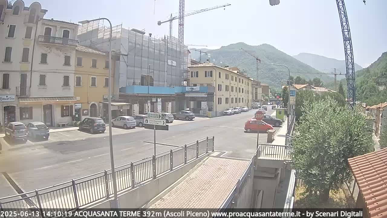 A sunny view of a town square with parked cars, buildings under construction, and mountains in the background.