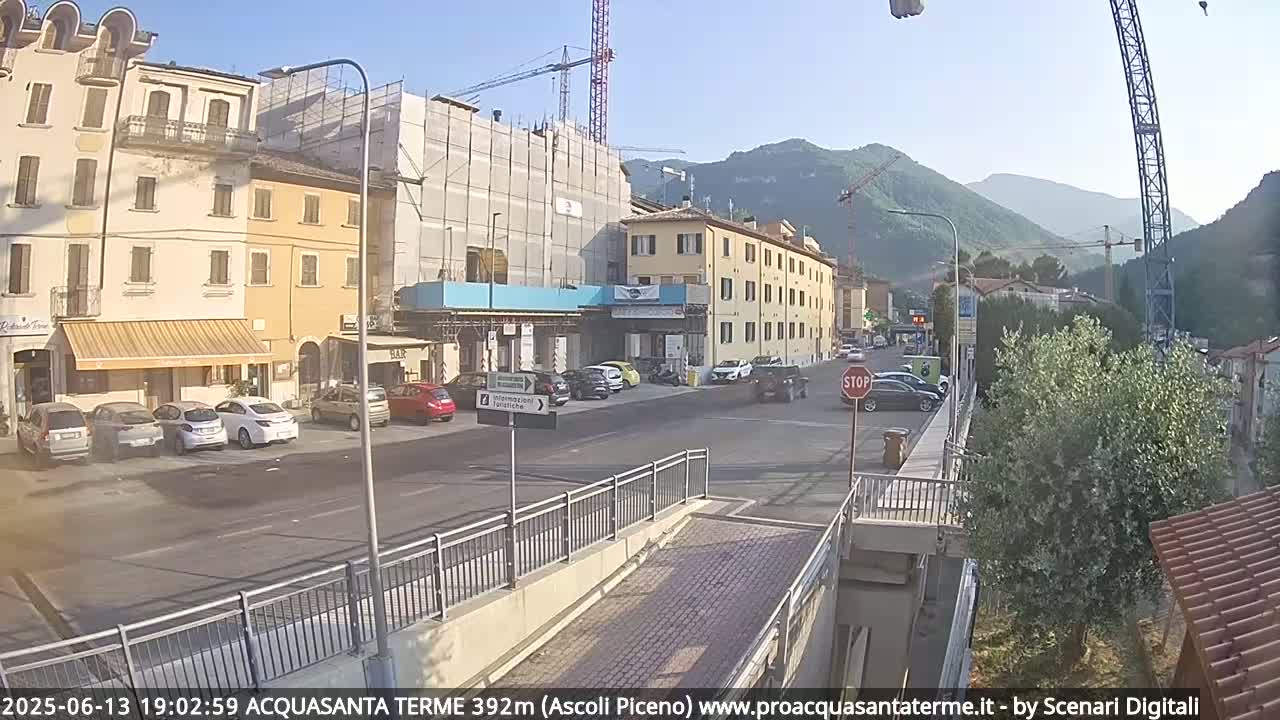 A sunny street scene in a town nestled in mountains, showing parked cars, buildings under construction, and some traffic.