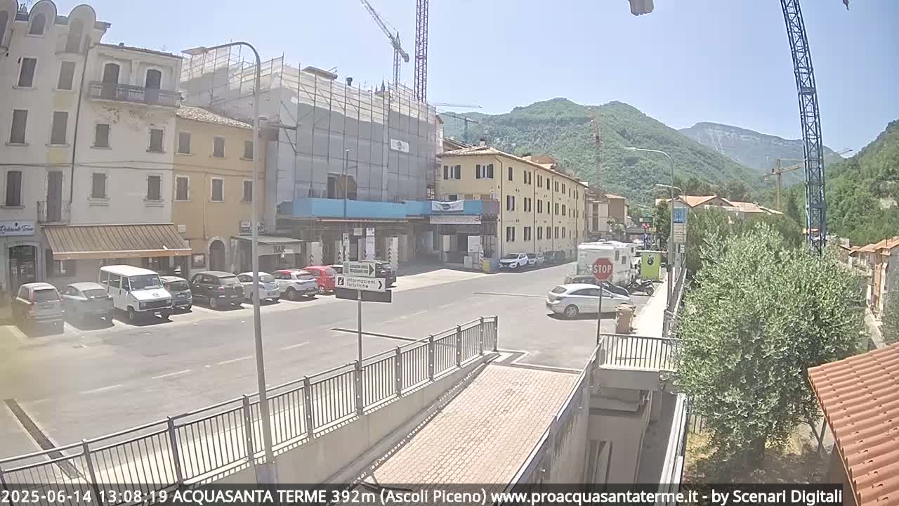 A sunny day reveals a town square with parked cars, buildings under construction, and a mountain range in the background.