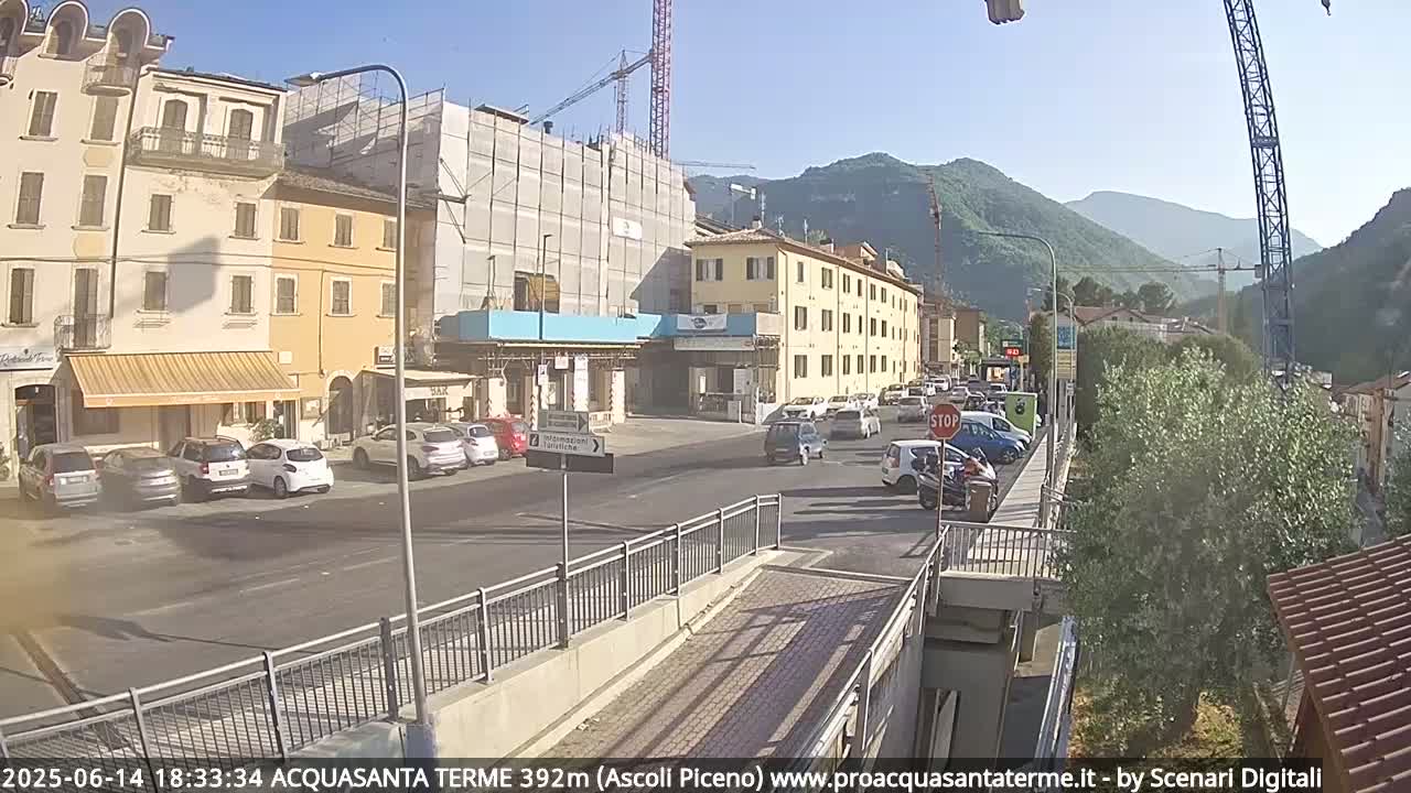 A sunny day in a town shows a street with parked and moving cars, buildings under construction, and mountains in the background.