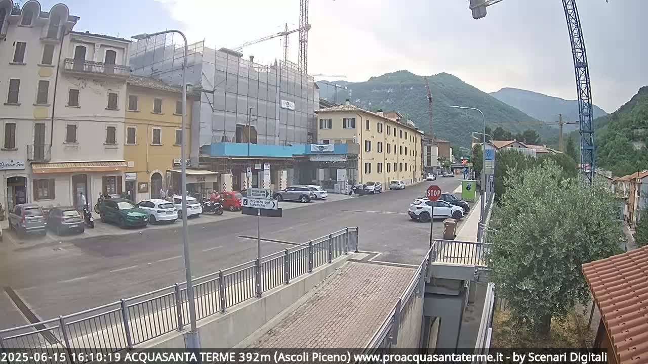 A street scene in a town nestled in mountains under a mostly clear sky, with cars parked along the side of the road and construction scaffolding visible on a building.