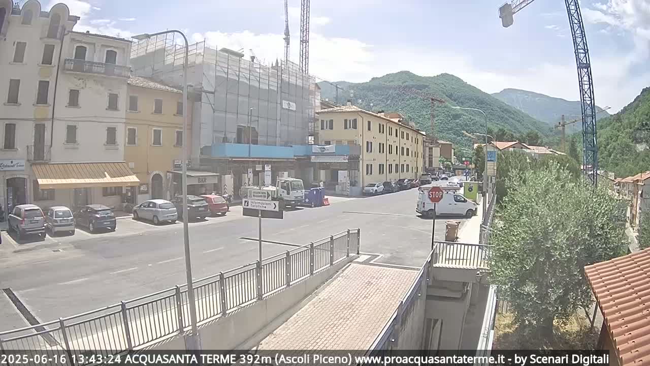 A sunny day reveals a town square with parked cars, buildings under construction, and mountains in the background.