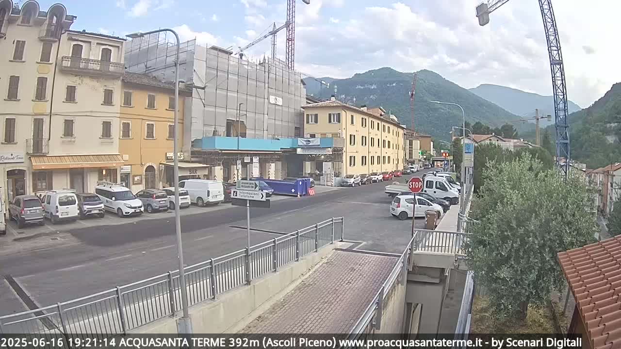 A partly cloudy day reveals a street scene in a town nestled in a valley between mountains, with parked cars lining the side of a road next to buildings, one of which is under construction.