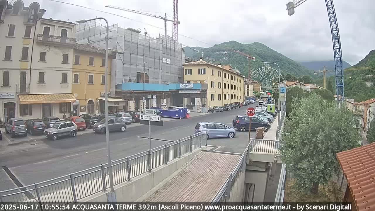 A street scene on an overcast day shows parked cars along the sides of a road in front of multi-story buildings, one of which is under construction, with hills in the background.