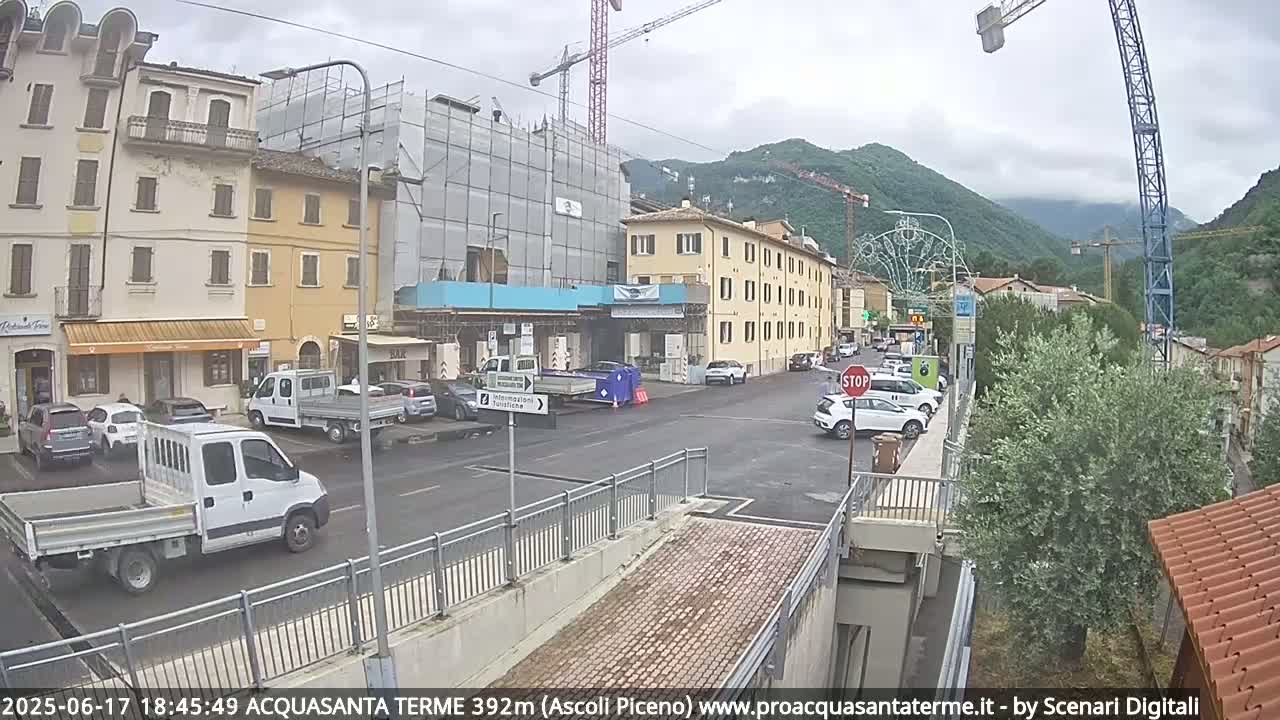 A street scene shows several cars parked in a lot next to multi-story buildings under a cloudy sky, with construction scaffolding visible on one building and mountains in the background.