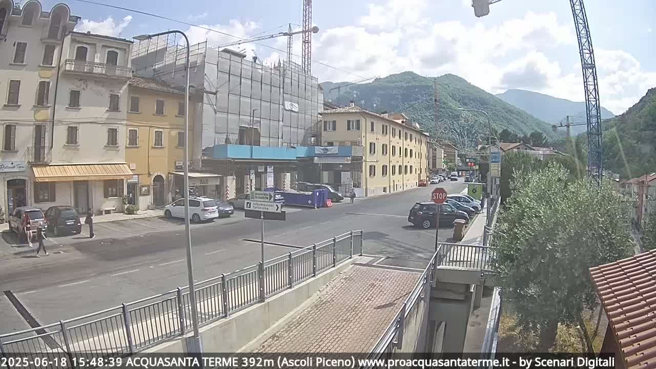 A sunny day reveals a town scene with parked cars, pedestrians, and buildings, including one under construction, all situated against a backdrop of mountains.