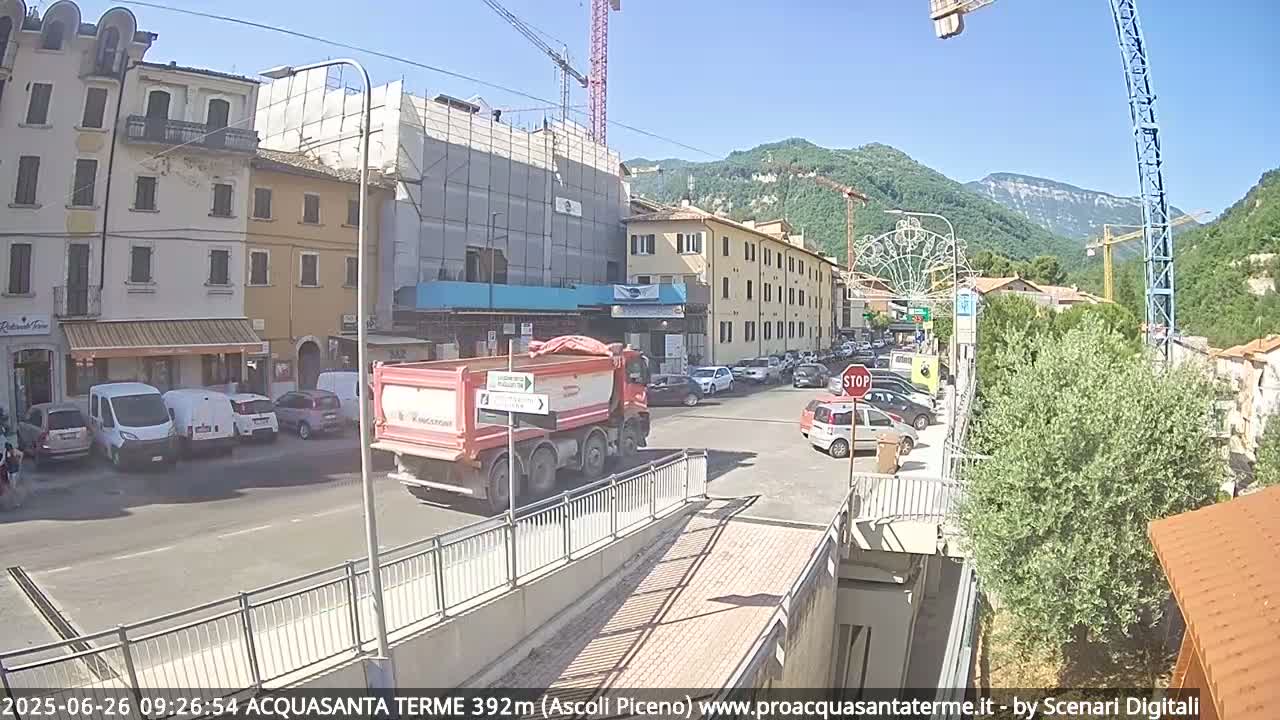 A sunny day reveals a town square with parked cars, a red dump truck, several buildings under construction, and mountains in the background.
