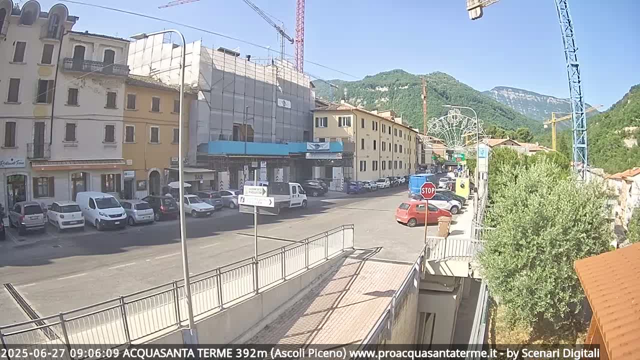 A sunny street scene shows several parked cars in front of multi-story buildings, one of which is under construction, with mountains visible in the background.