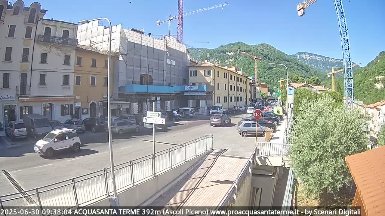 A sunny street scene shows cars parked and driving on a road between buildings under construction, with mountains in the background and a construction crane visible.