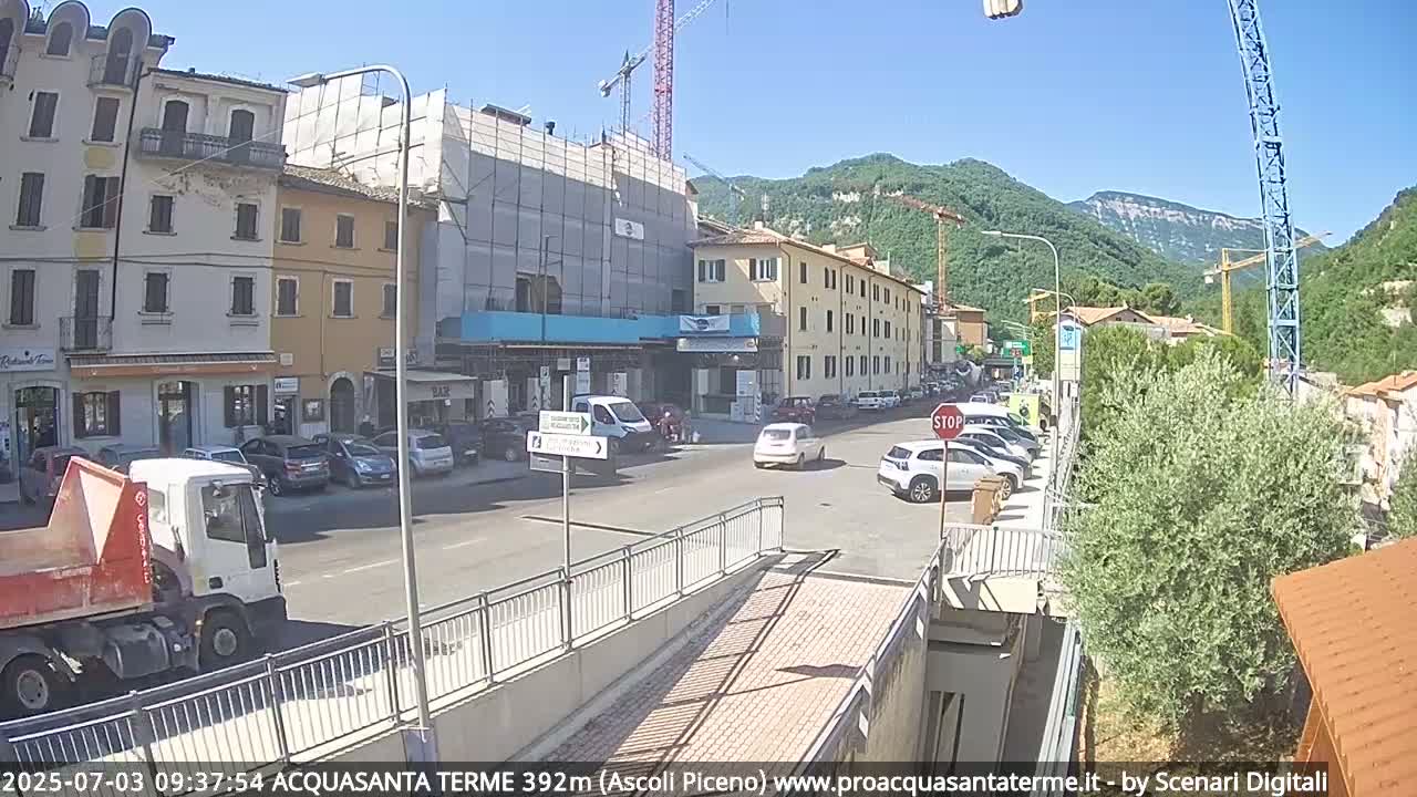 A sunny day reveals a town square with buildings under construction, parked cars, and a truck, all situated beside a hill.