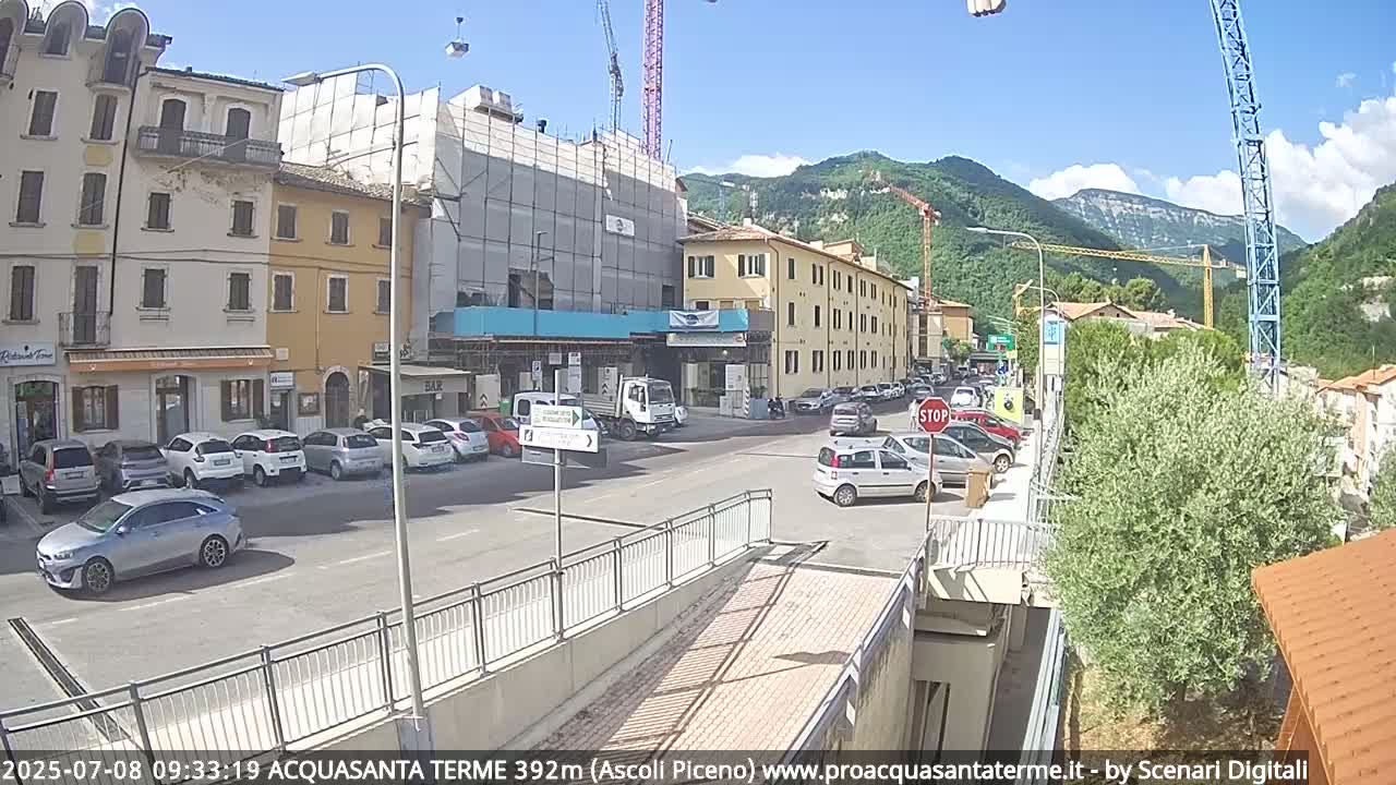 A sunny day in a town with several parked cars, buildings under construction, and mountains in the background.