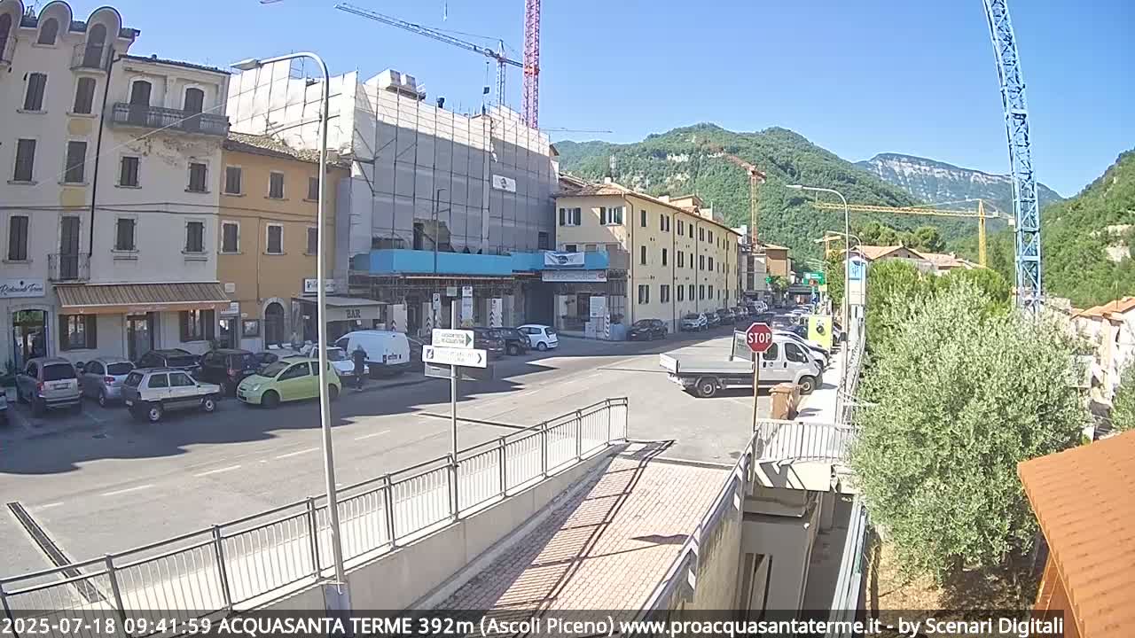 A sunny street scene shows several parked cars, buildings under construction, and a few pedestrians in a town nestled against a mountainside.