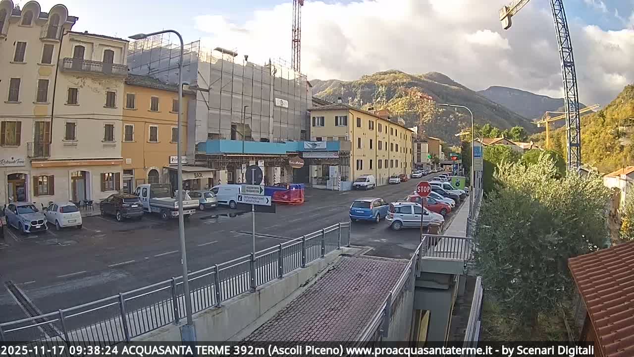 On a partly cloudy day, a street in a mountainous town is filled with parked cars and moving vehicles, flanked by multi-story buildings, including one covered in scaffolding, with two large construction cranes prominent against a backdrop of green and autumn-hued hills.