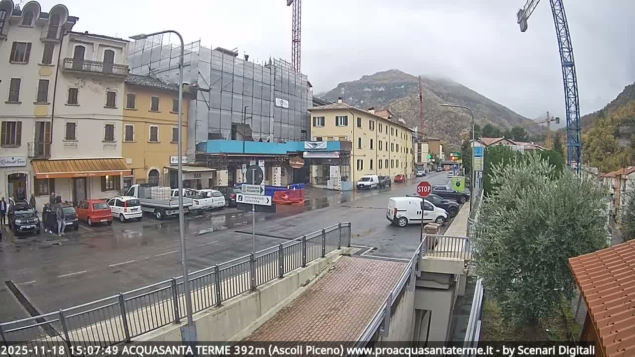On a partly cloudy day, a street in a mountainous town is filled with parked cars and moving vehicles, flanked by multi-story buildings, including one covered in scaffolding, with two large construction cranes prominent against a backdrop of green and autumn-hued hills.