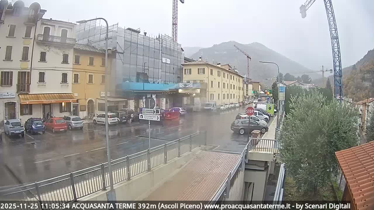 A wide-angle view captures a rainy, overcast day in a European town, with wet streets lined by multi-story buildings, numerous parked cars, active construction featuring large cranes, and hazy hills in the background.