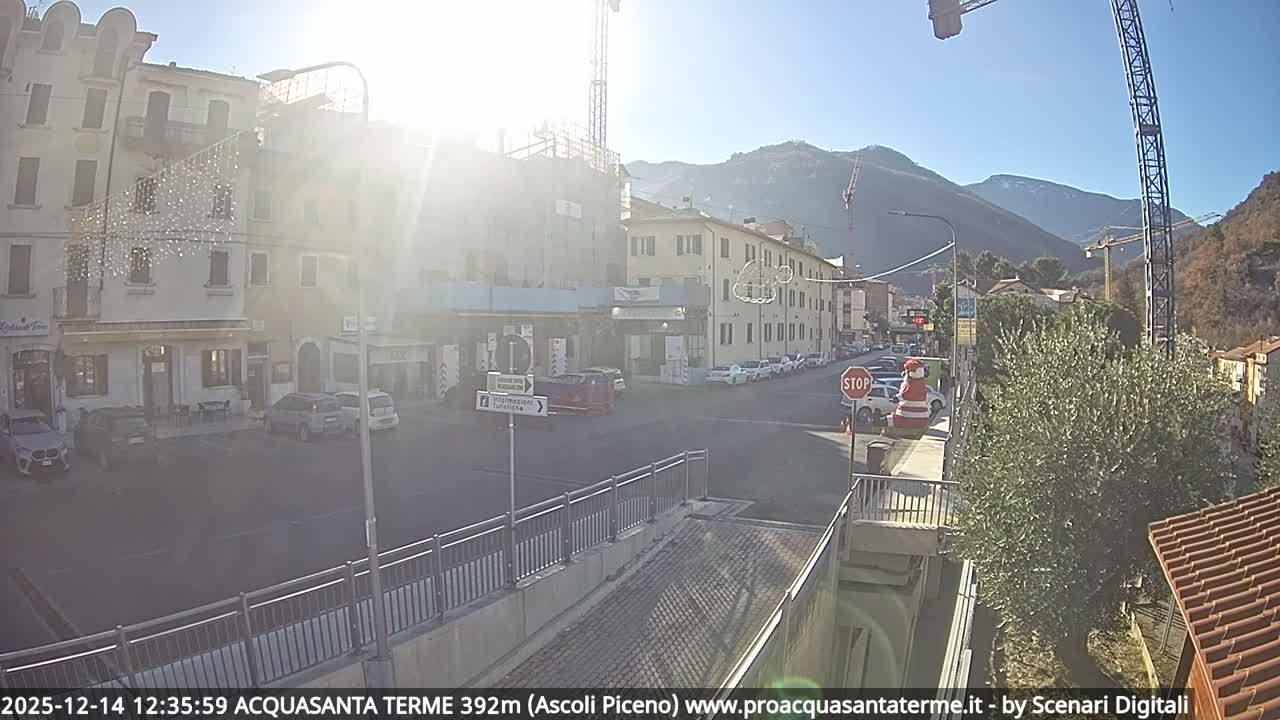 A wide-angle view captures a rainy, overcast day in a European town, with wet streets lined by multi-story buildings, numerous parked cars, active construction featuring large cranes, and hazy hills in the background.
