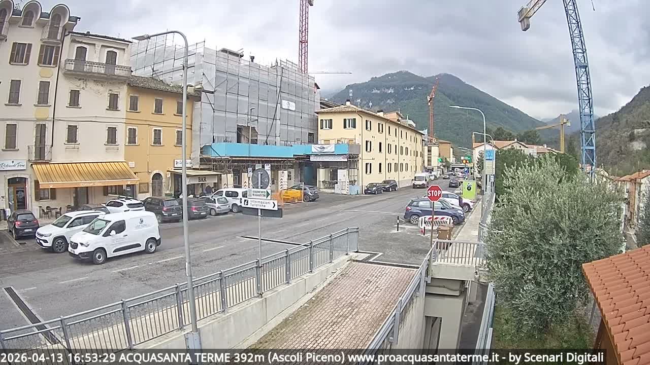 A wide-angle view captures a rainy, overcast day in a European town, with wet streets lined by multi-story buildings, numerous parked cars, active construction featuring large cranes, and hazy hills in the background.
