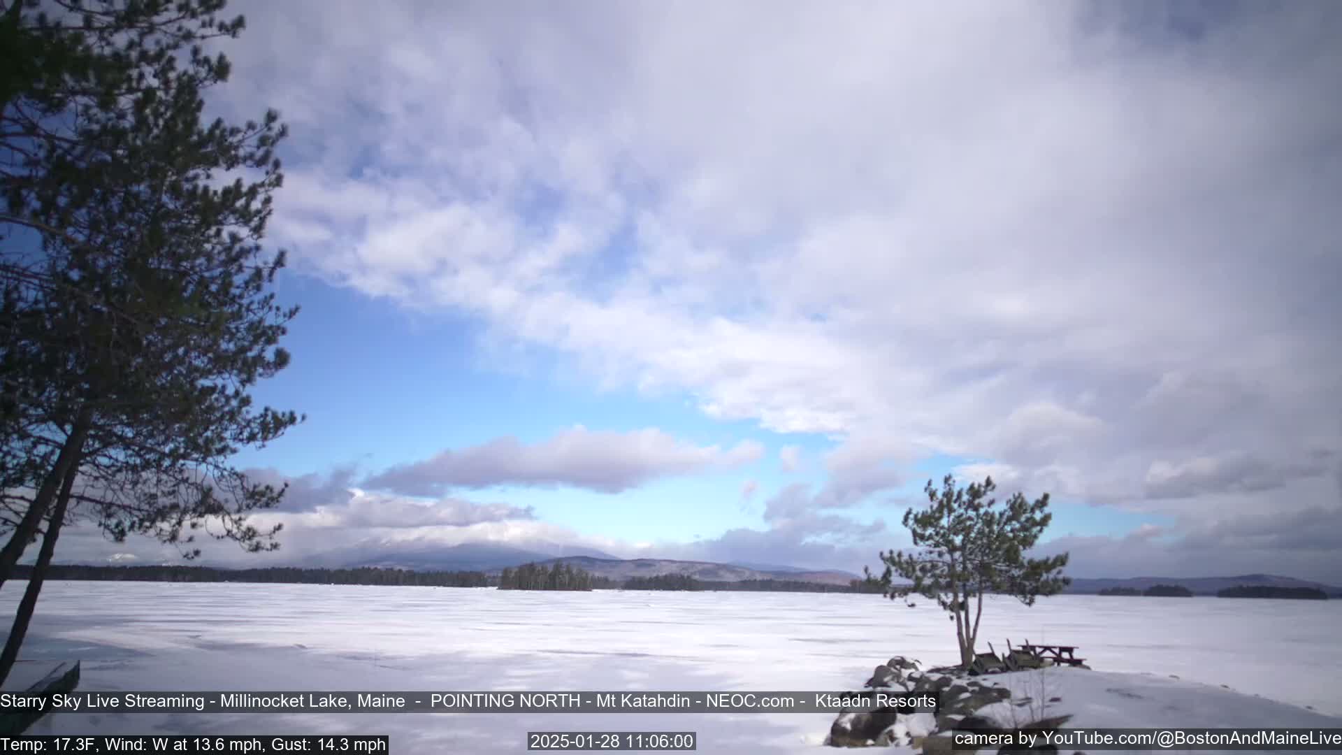 A snow-covered lake stretches to distant mountains under a partly cloudy sky, with a few trees in the foreground.