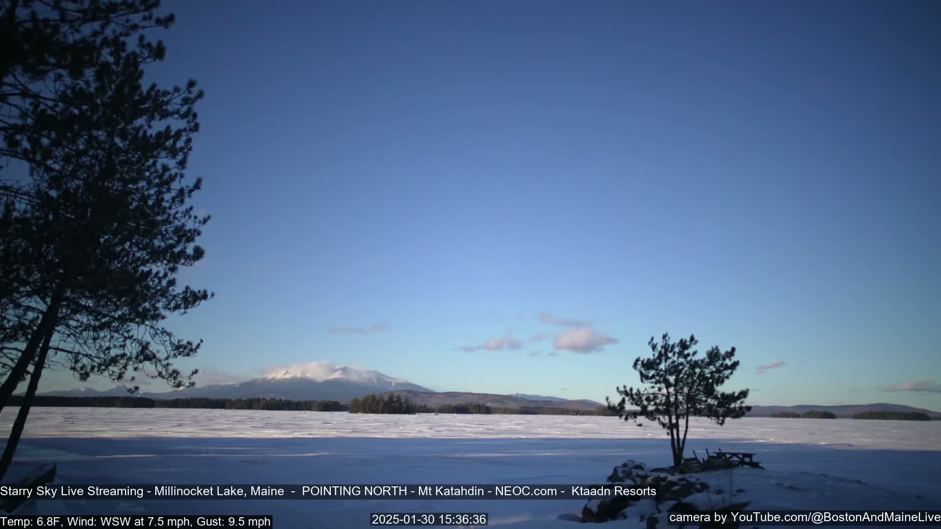 A snow-covered lake stretches to a distant mountain range under a mostly clear, blue sky.