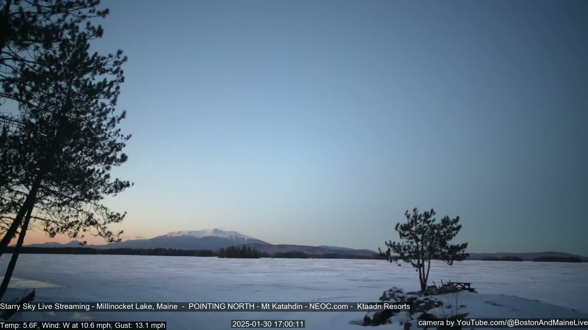 A frozen lake under a clear, twilight sky, with silhouetted trees in the foreground and distant mountains on the horizon.