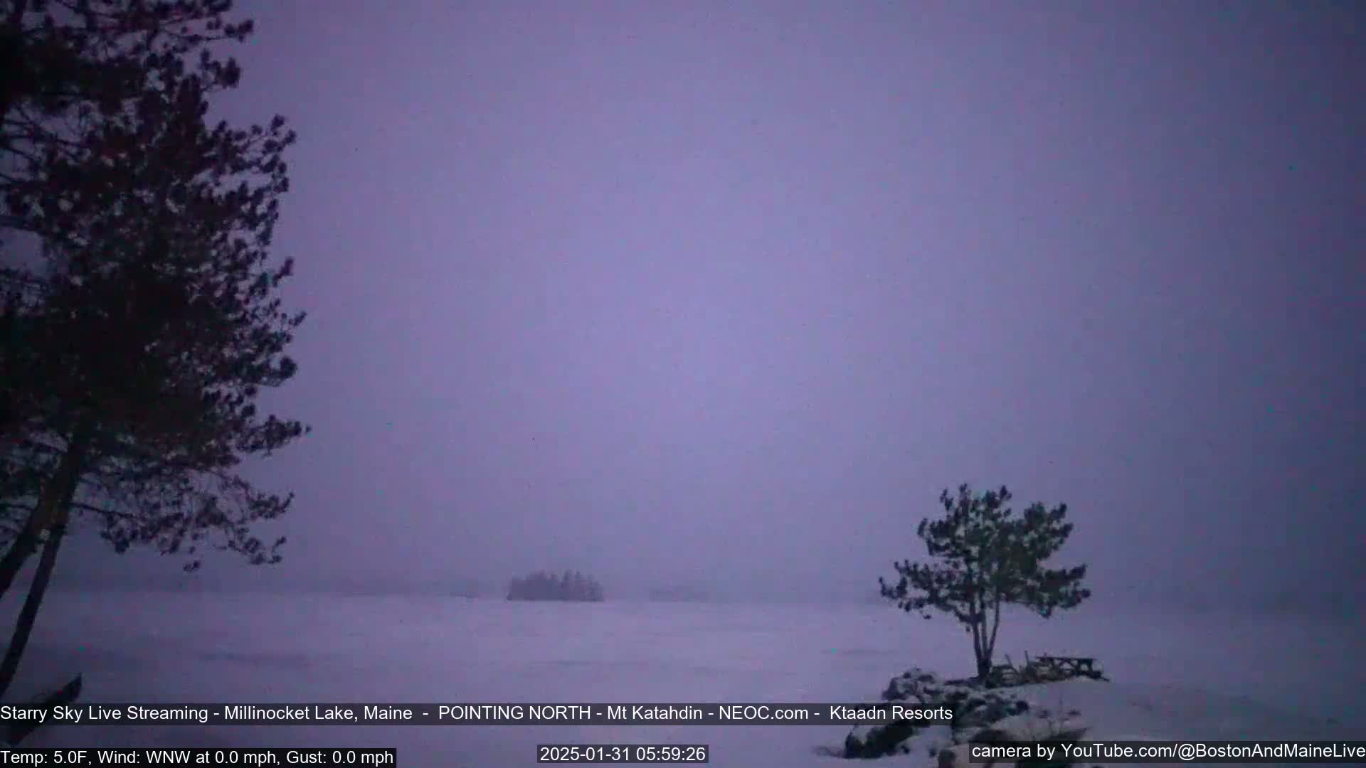 A snowy, hazy landscape shows a lake partially obscured by mist with dark silhouettes of trees and a small island in the distance under a dusky sky.