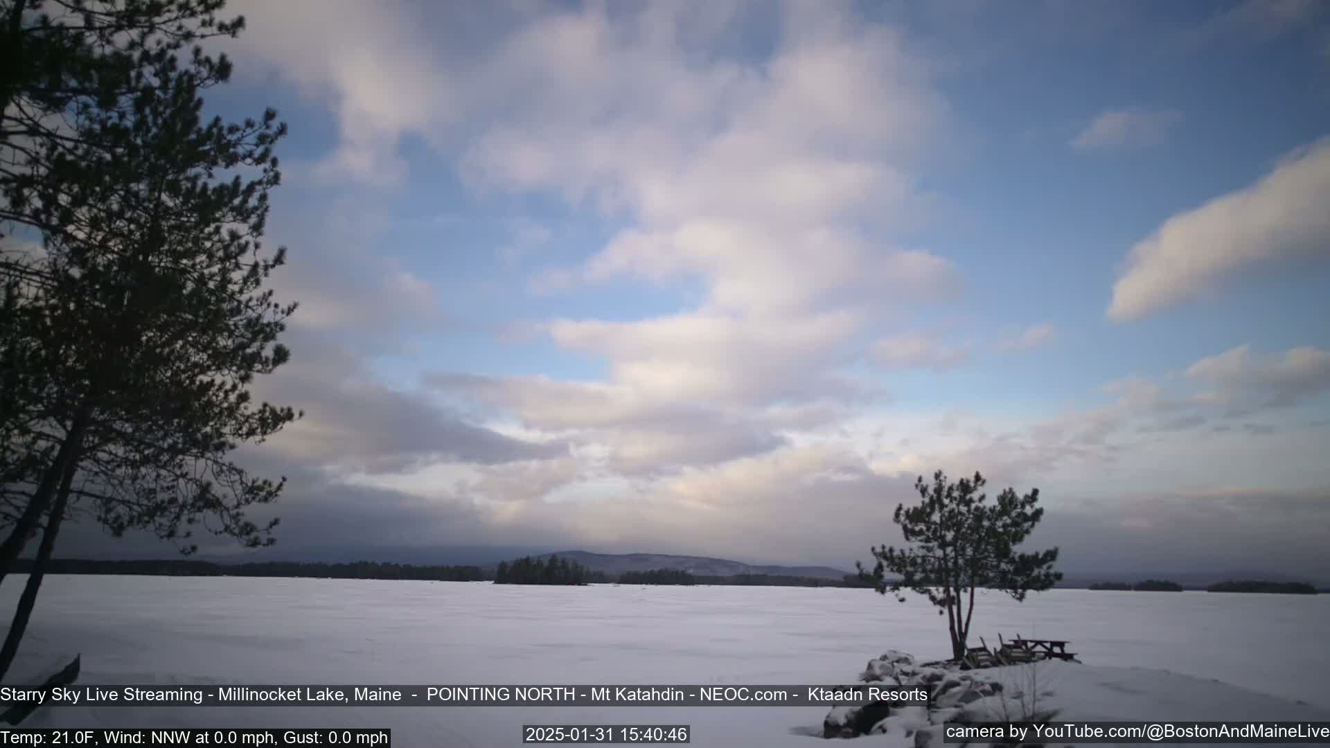 A snow-covered lake stretches to a distant, tree-lined shore under a partly cloudy, pale blue sky.