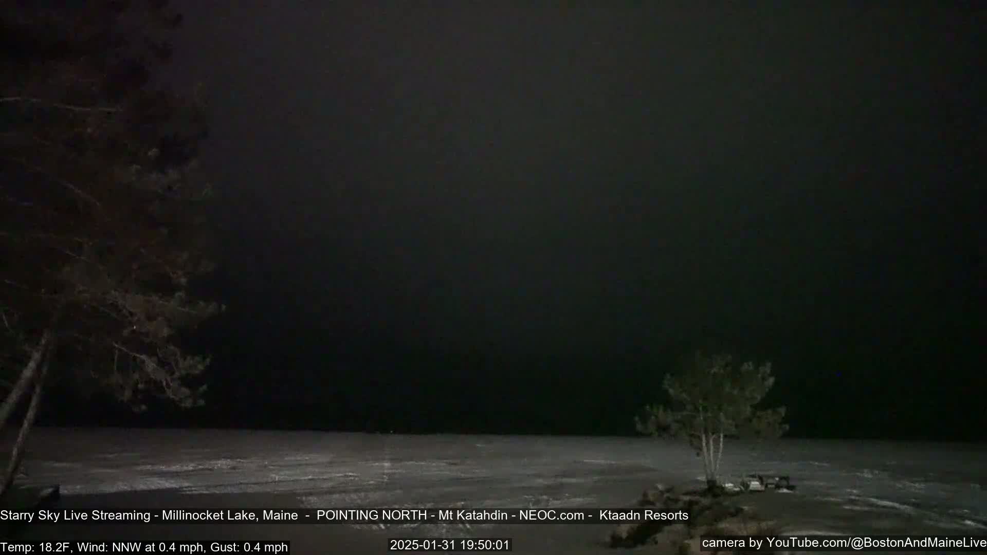 A dark, nighttime scene shows a mostly frozen lake under a cloudy sky, with a few trees visible near the shoreline.