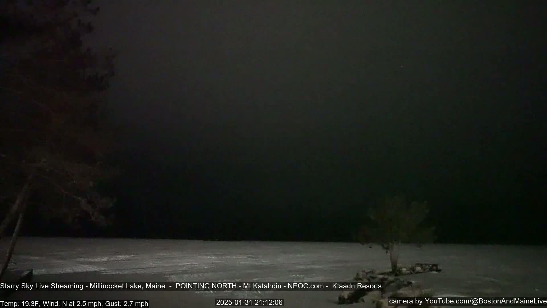A dark, nighttime scene shows a mostly frozen lake under a nearly cloudless sky, with a few bare trees visible on the shore.