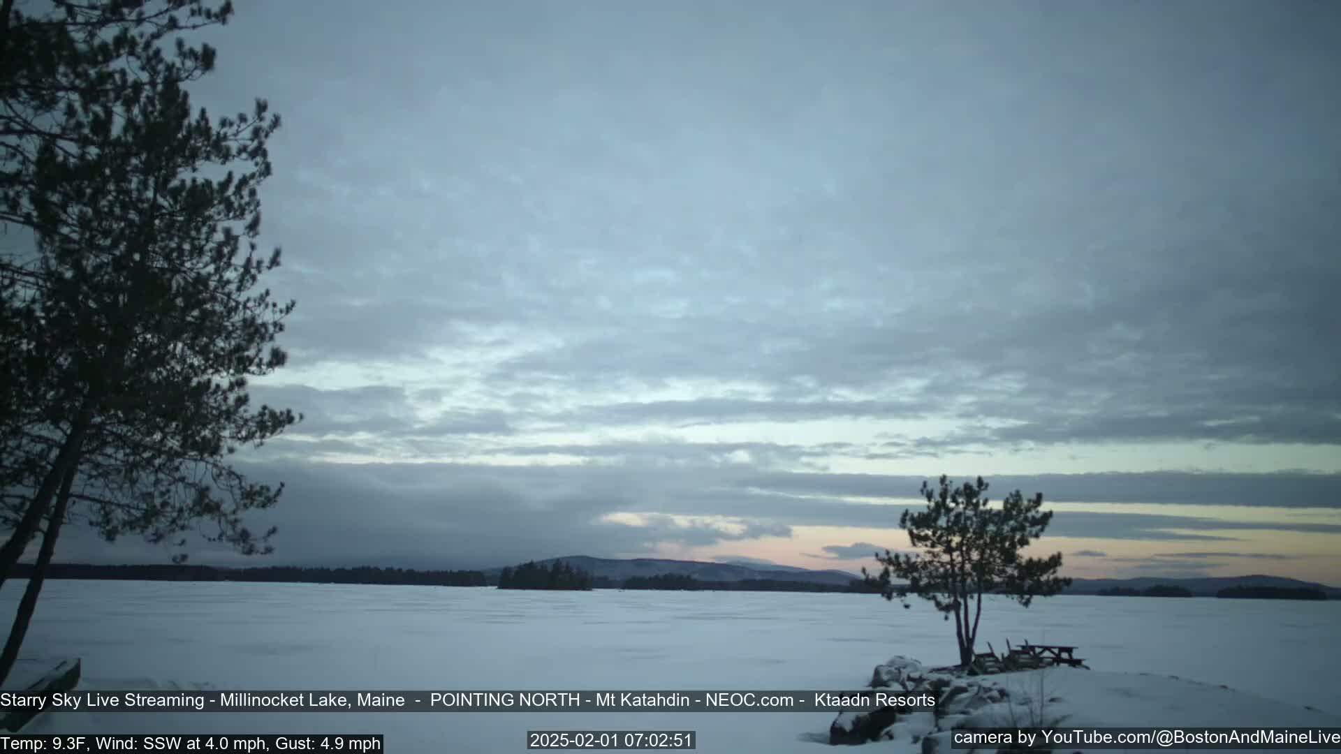 A snow-covered lake is seen at dawn under a cloudy sky with a few distant trees and mountains visible in the background.
