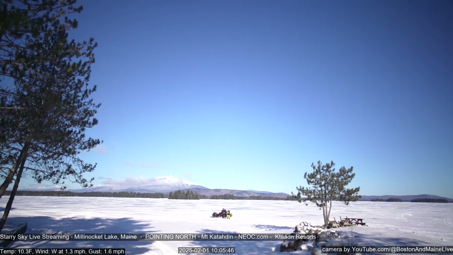 A snow-covered lake stretches to distant mountains under a clear blue sky, with a snowmobile and its riders visible in the middle distance.