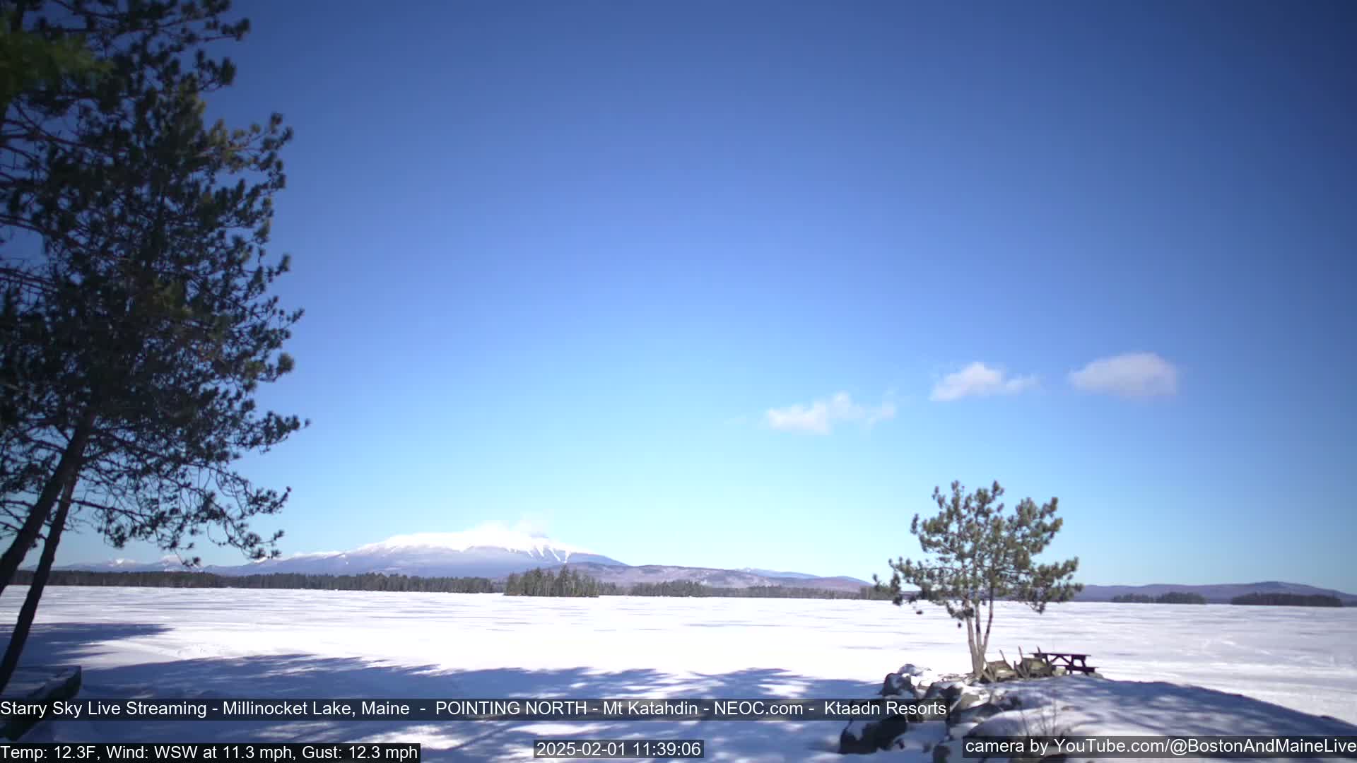 A snow-covered lake stretches to a mountain range under a mostly clear, bright blue sky.