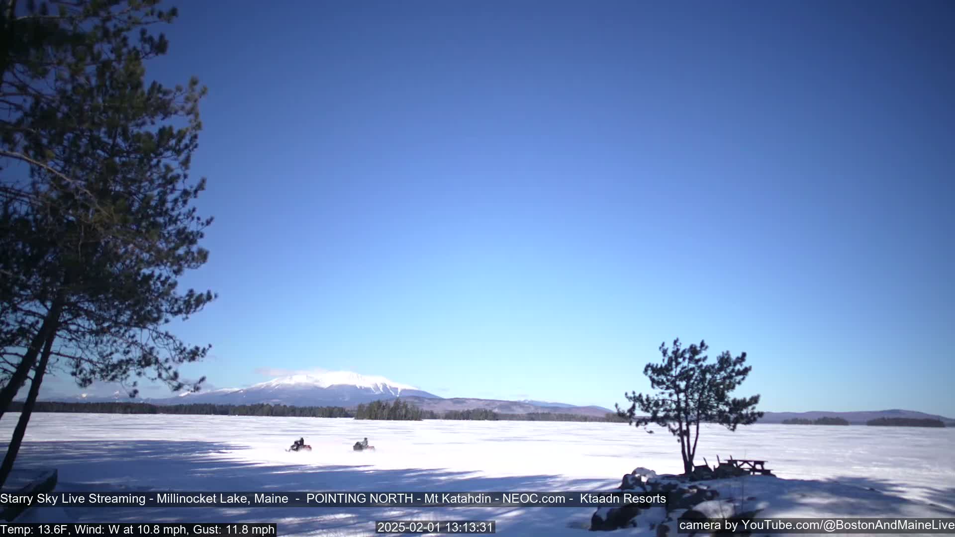 Two snowmobiles travel across a frozen lake under a clear blue sky, with a snow-covered mountain range in the distance.