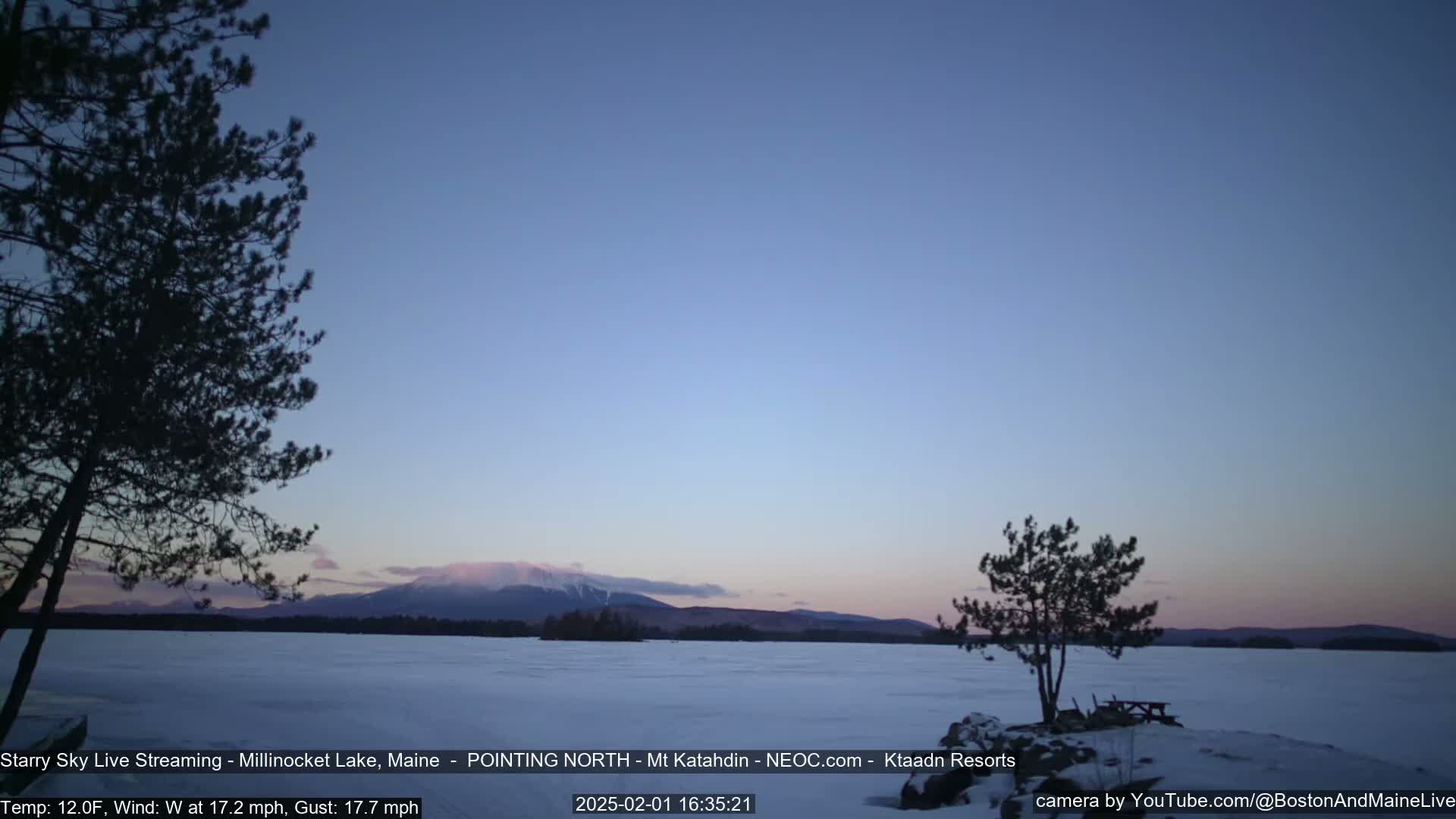 A snow-covered lake with silhouetted trees in the foreground and a mountain range in the background under a clear, twilight sky.