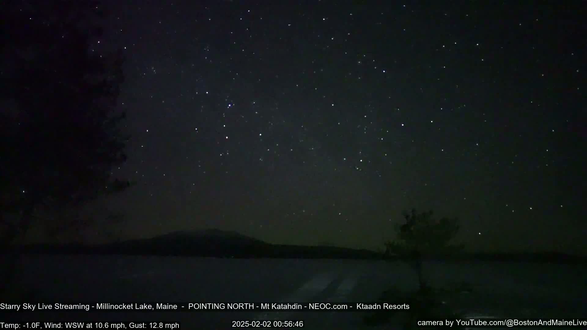 A nighttime long exposure shows a dark lake with a distant mountain range under a clear, starry sky with a slight wind.