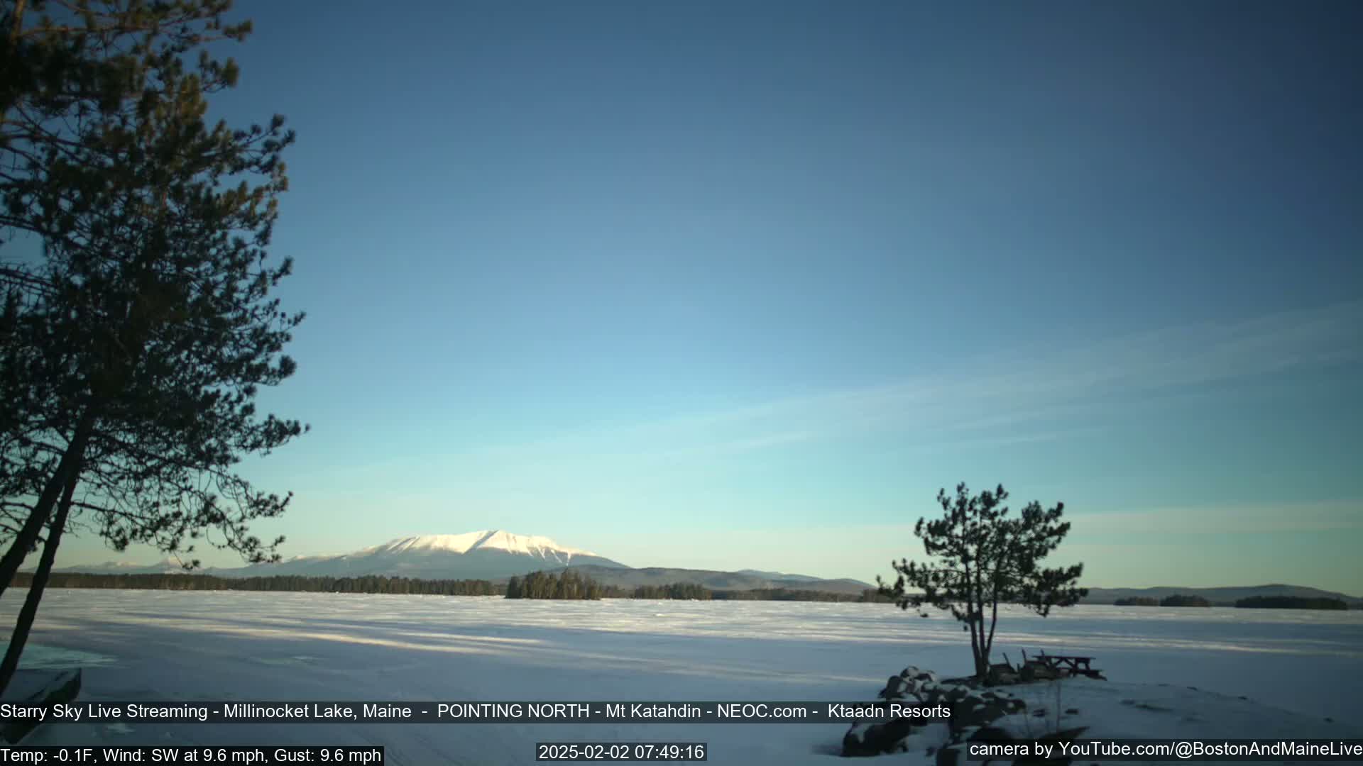 A snow-covered lake stretches to a distant mountain range under a clear blue sky, with a few trees in the foreground.