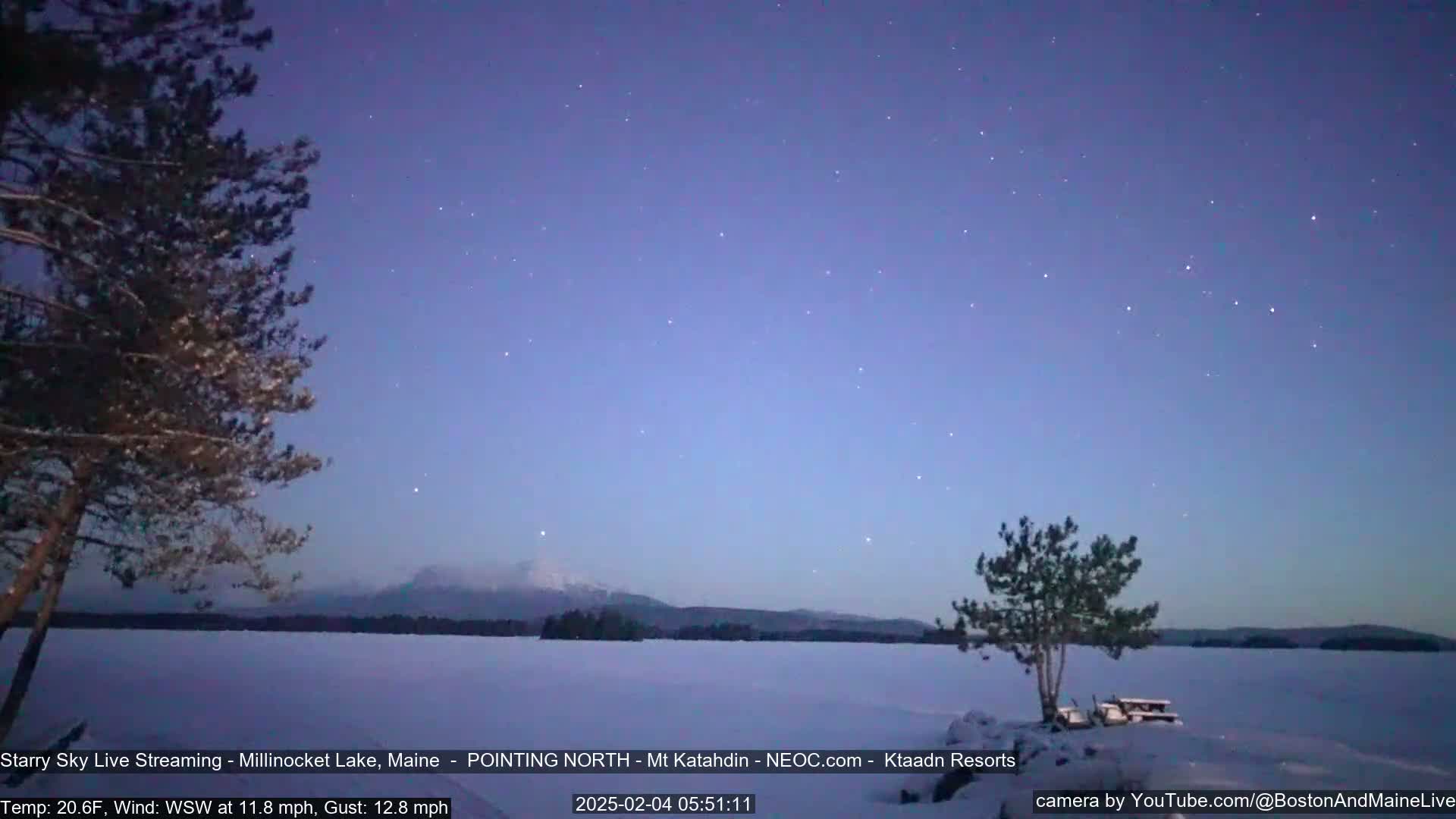 A snow-covered lake is seen at twilight under a starry sky, with a distant mountain range visible on the horizon and sparsely wooded shorelines.